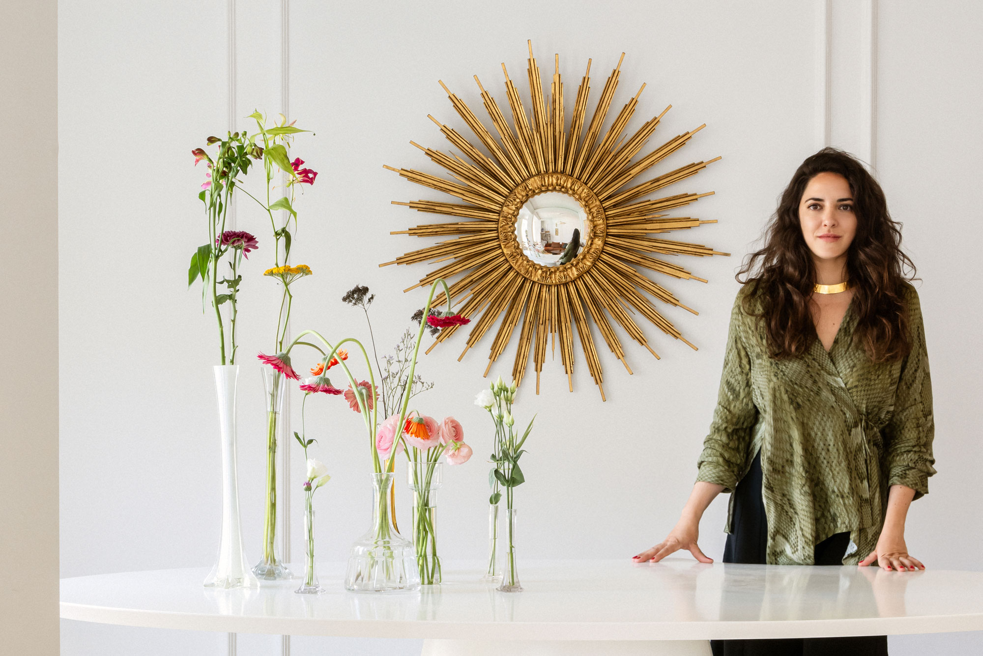 Mujer diseñadora de interiores junto a una mesa con flores, frente a un espejo dorado de sol en un espacio luminoso y elegante.